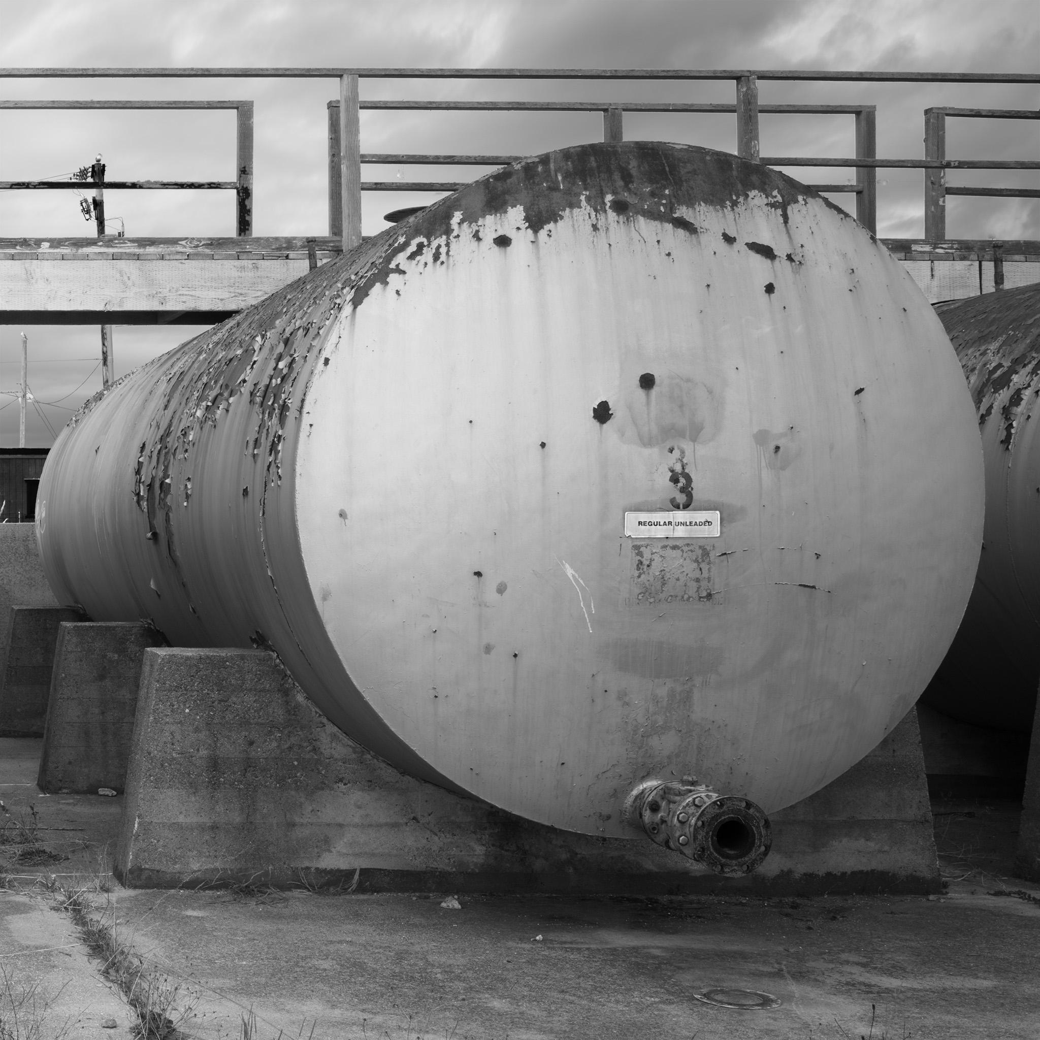 A large, cylindrical tank with peeling paint, on concrete blocks, stenciled with the digit "3" on the end with a label that says "Regular Unleaded" below. A spigot protrudes from the tank at the bottom on the end.
