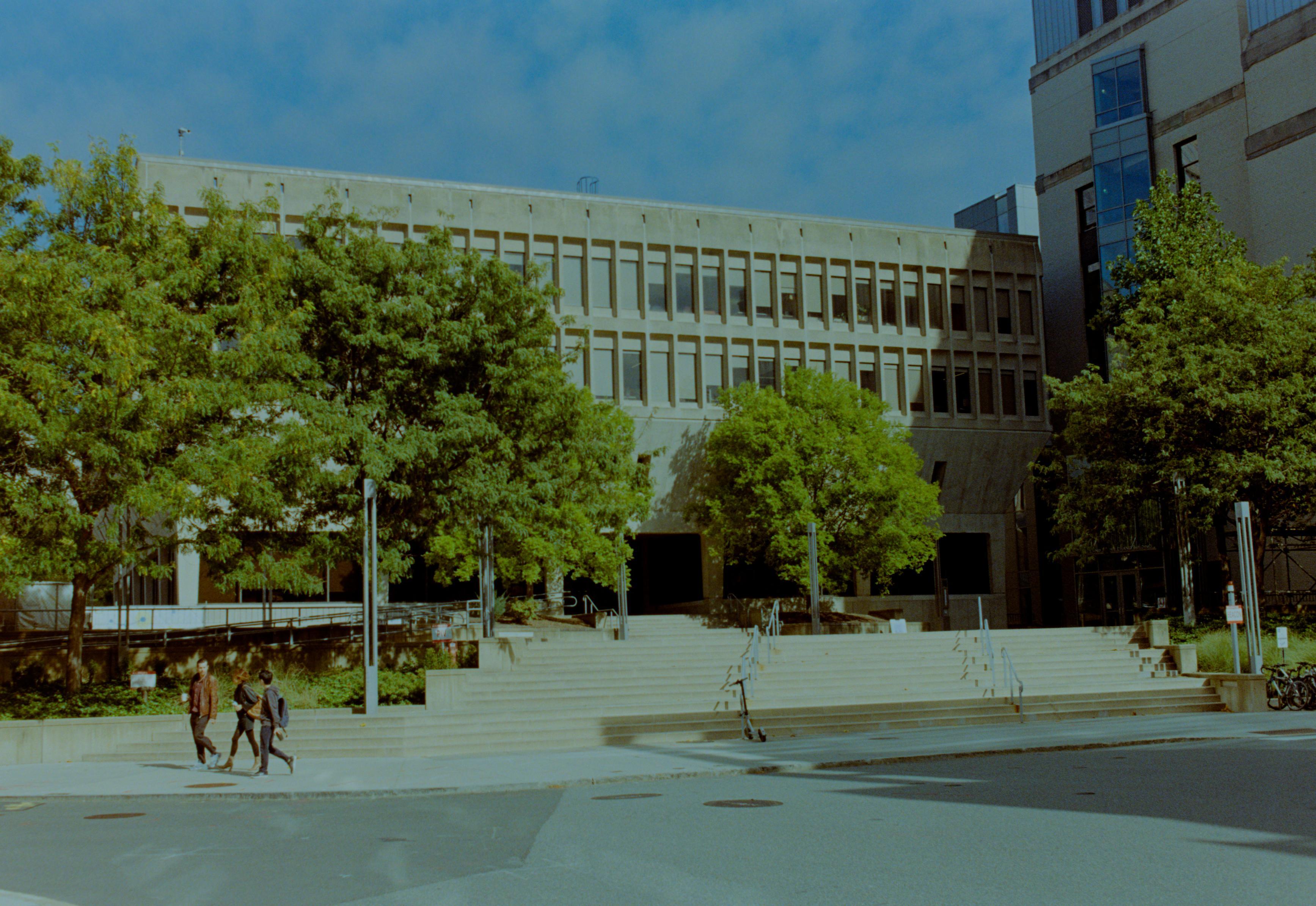 People walk outside a brutal stone building. Summer reigns.