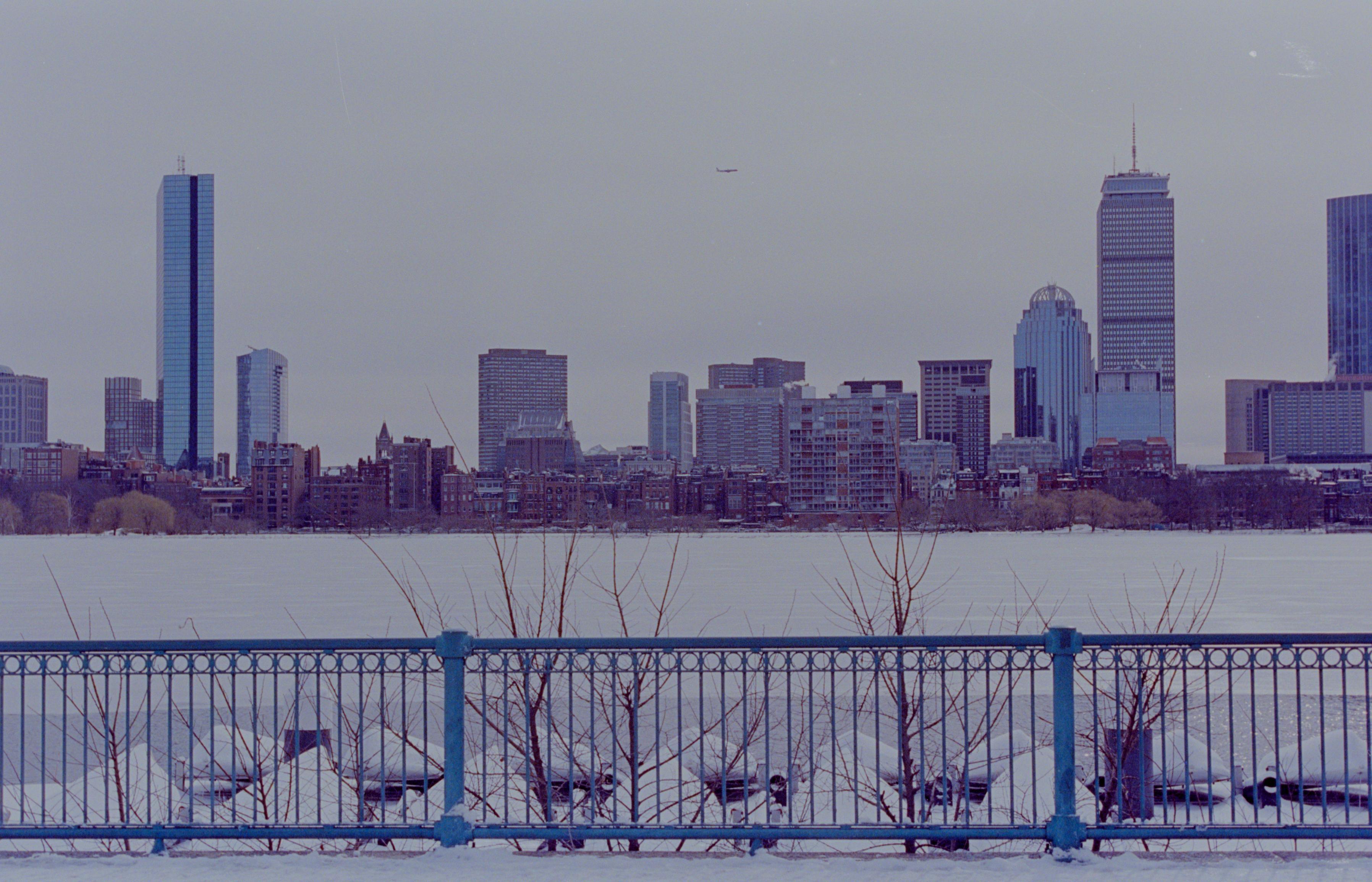 Boston skyline seen from across the Charles river. The river is frozen over. A plane sits in the sky framed by the buildings in the distance. In the foreground, a blue metal fence protects the people from the icy water below. 