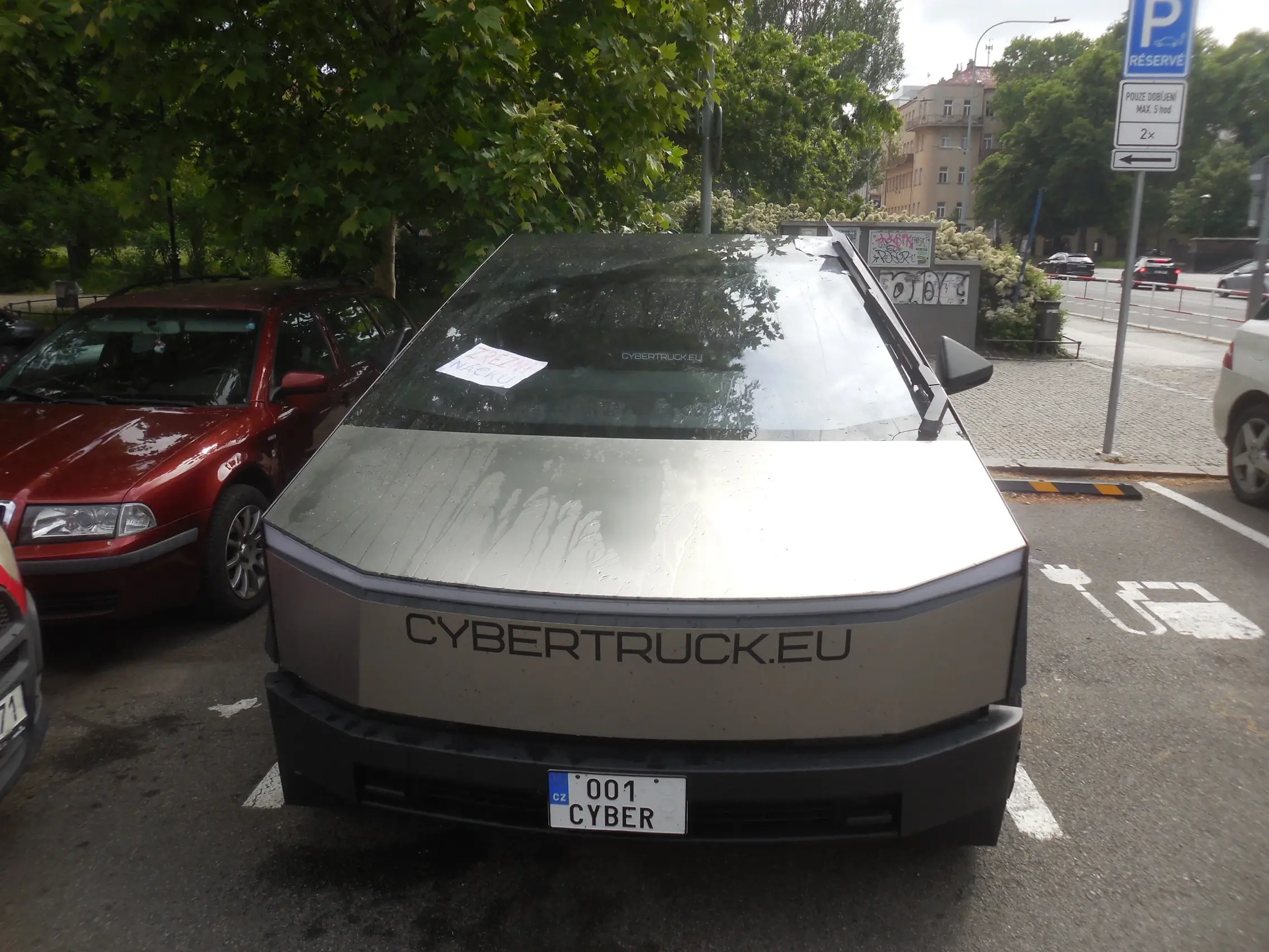 Tesla Cybertruck with custom near-invisibly rounded edges to pass EU standards, charging at a parking lot in central Prague. It is monstrous compared to a 2000s family car next to it. Giant "CYBERTRUCK.EU" sticker on the front. Licence plate: "001 CYBER". The weather is sunny but the car has been splashed with water, with a sheet of paper left on the windshield.