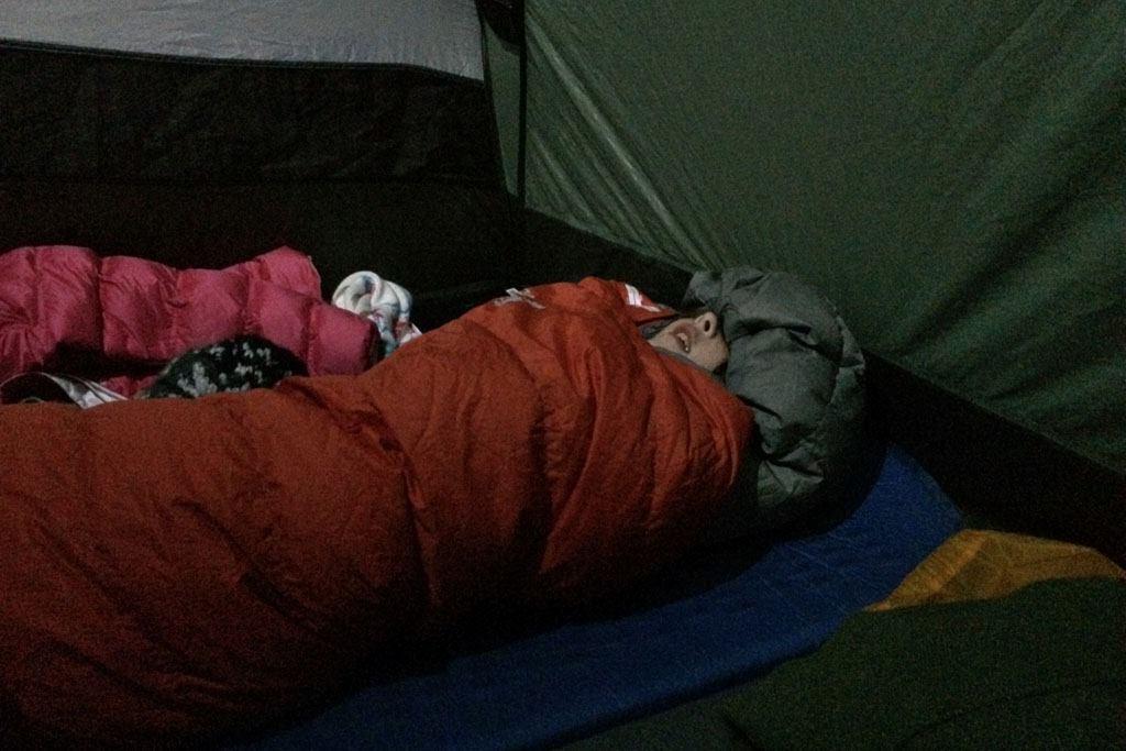 Inside a tent a portion of a kid's face peeks out from the hood of a red sleeping bag.